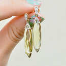 Gold and quartz earrings held by a hand with a floral nail design on a light background